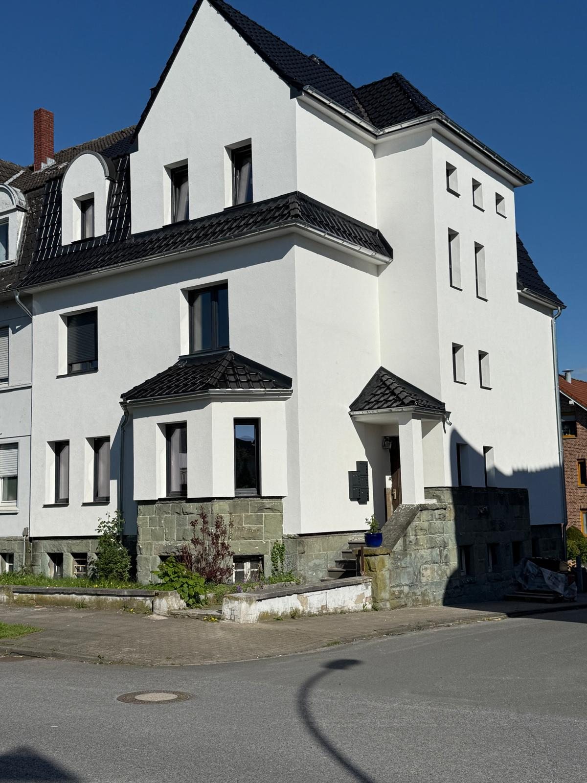 Weißes, mehrgeschossiges Stadthaus mit Erker, dunklem Dach und dunkel gerahmten Fenstern vor blauem Himmel
